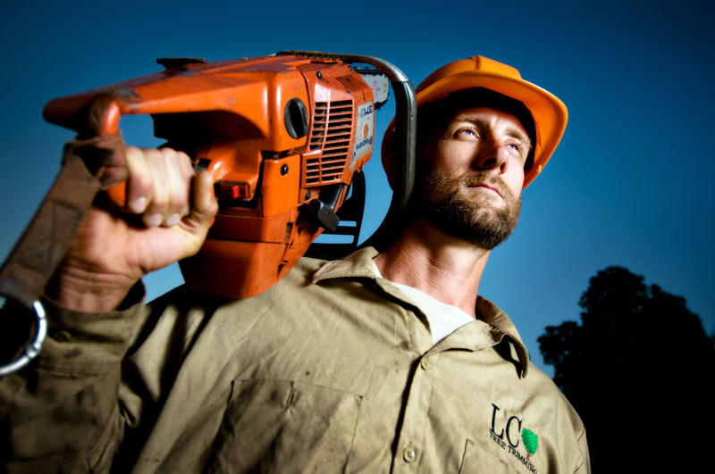 Construction worker in a yellow hard hat carries an orange chainsaw on his shoulder against a blue sky.